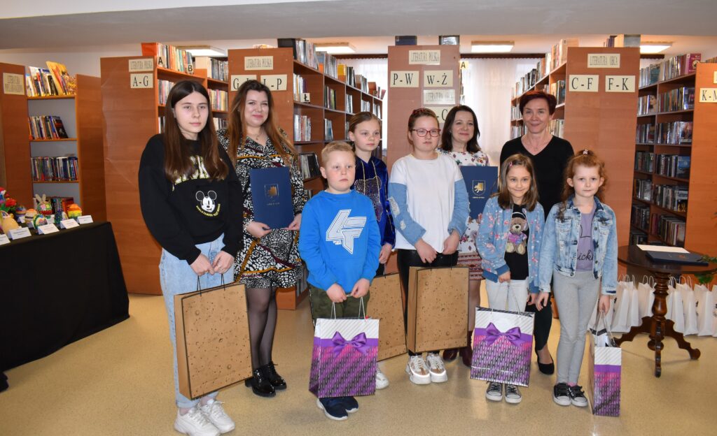 Grupa zwycięzców z konkursu na "Najpiękniejszą pisankę w Gminie Dobre" stoją z nagrodami. W tle regały biblioteczne z książkami, stół z pisankami.