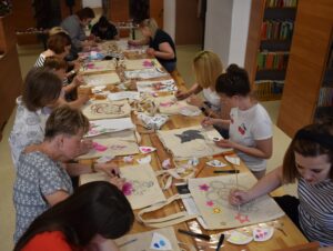 Grupa kobiet ze skupieniem maluje swoje torby kolorowymi farbami, za nimi regał z książkami