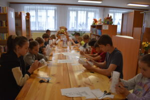 Przy drewnianym stole dzieci siedzą i rysują na szklanych wazonach. W tle regały z kolorowymi książkami
