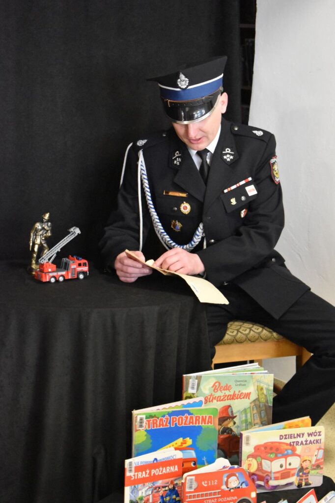 Strażak w mundurze czyta książkę na stole stoi samochód strażacki i statuetka strażaka, na dole stoją książki o strażakach.