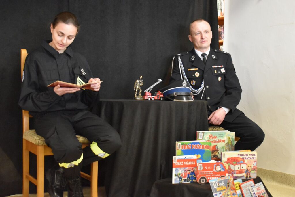 Przy czarnym stole siedzi pan i pani strażak która czyta książkę. Na stole leży czapka, samochód i statuetka strażacka. Na dole stoją książki o strażakach, wąż i sikawka.