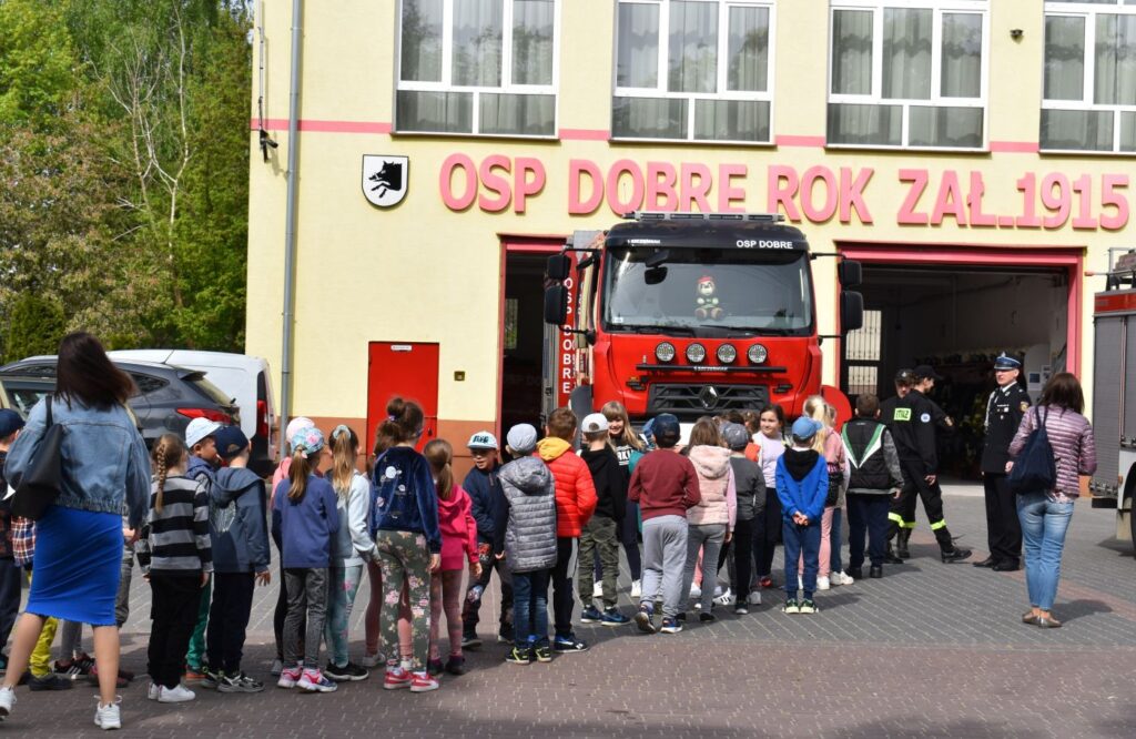 Grupa dzieci ustawiona w parach przyszła do jednostki Ochotniczej Straży Pożarnej w Dobrem. Przed budynkiem stoi czerwony wóz strażacki i troje strażaków.