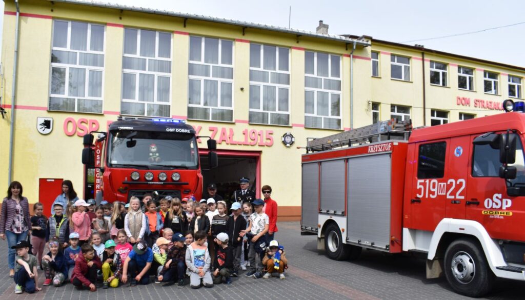 Przed budynkiem Ochotniczej Straży Pożarnej stoją dwa wozy strażackie i grupka dzieci z paniami i strażakami pozują do zdjęcia