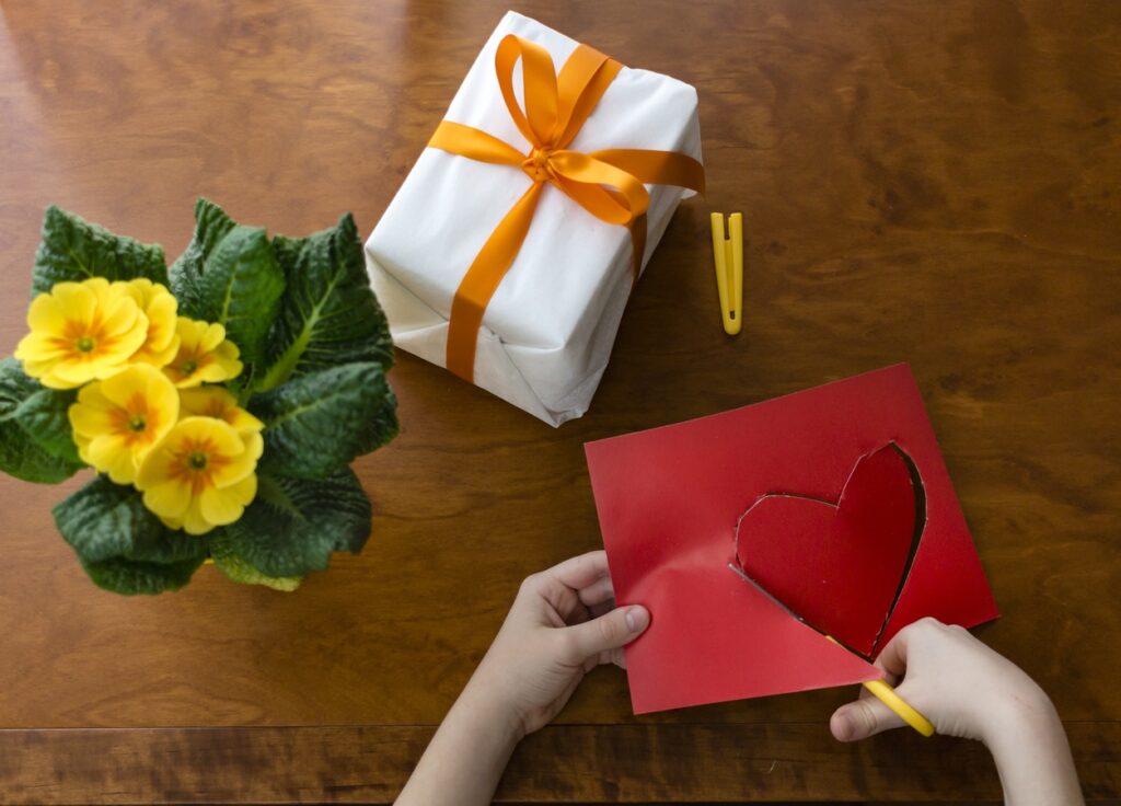 widok z góry na kwiatek doniczkowy, zapakowany prezent i ręce wycinające nożyczkami serce z papieru