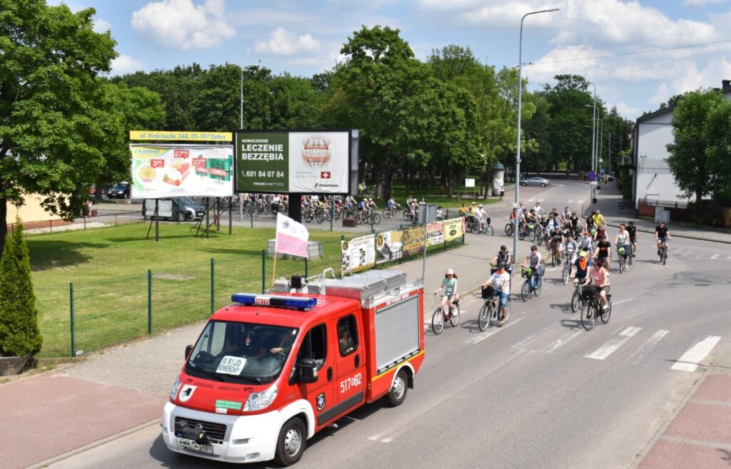 Czerwony wóz strażacki za nim dużo rowerzystów na skrzyżowaniu w tle park