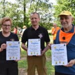 Przedstawiciele Ochotniczych Straży Pożarnych z dyplomami za udział w rajdzie rowerowym. W tle rowerzyści i wysokie zielone drzewa