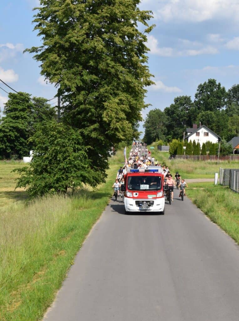Z górki zjeżdża peleton rowerzystów, prowadzi ich wóz strażacki. Z boku rośnie wysokie drzewo, w tle stoi biały dom