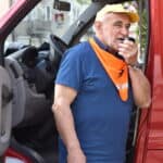 Przez mikrofon mówi mężczyzna w żółtej czapce i pomarańczowej chuście. W tle szoferka samochodu strażackiego.