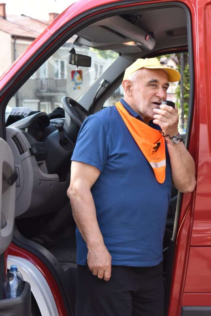 Przez mikrofon mówi mężczyzna w żółtej czapce i pomarańczowej chuście. W tle szoferka samochodu strażackiego.