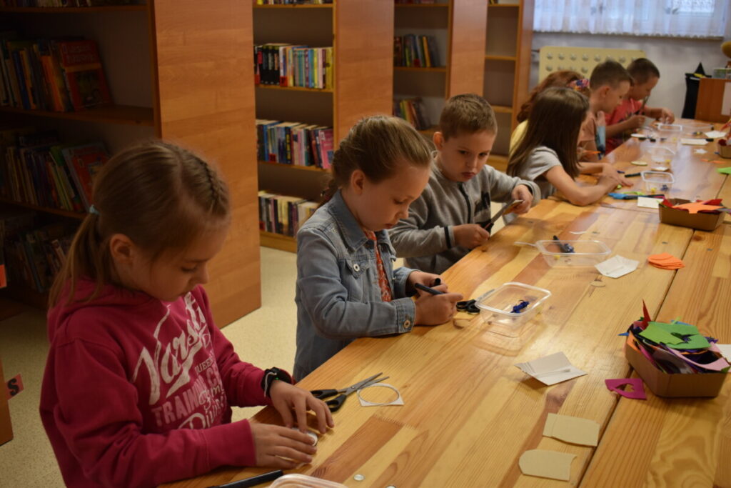 Dzieci siedzą przy drewnianym stole w Bibliotece,rysują i wycinają