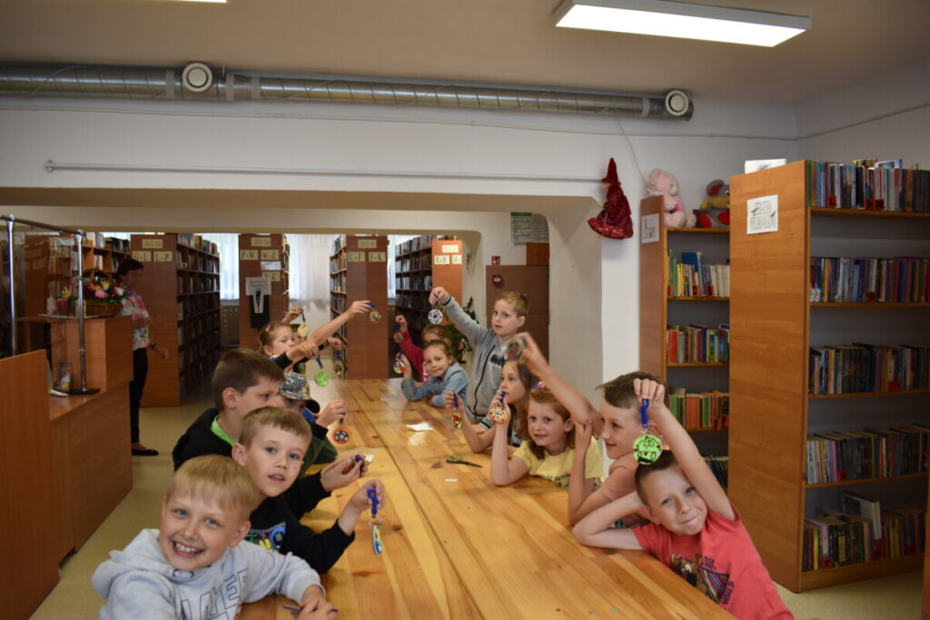 Dzieci siedzą przy stole i pokazują breloczki jakie zrobili dla swoich tatusiów. W tle regały biblioteczne.
