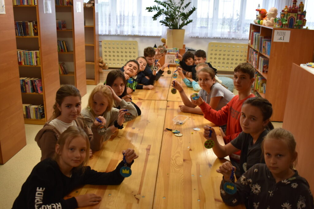 Dzieci siedzą przy stole w ręku trzymają okrągłe breloczki które zrobili samodzielnie. W tle regały biblioteczne z książkami