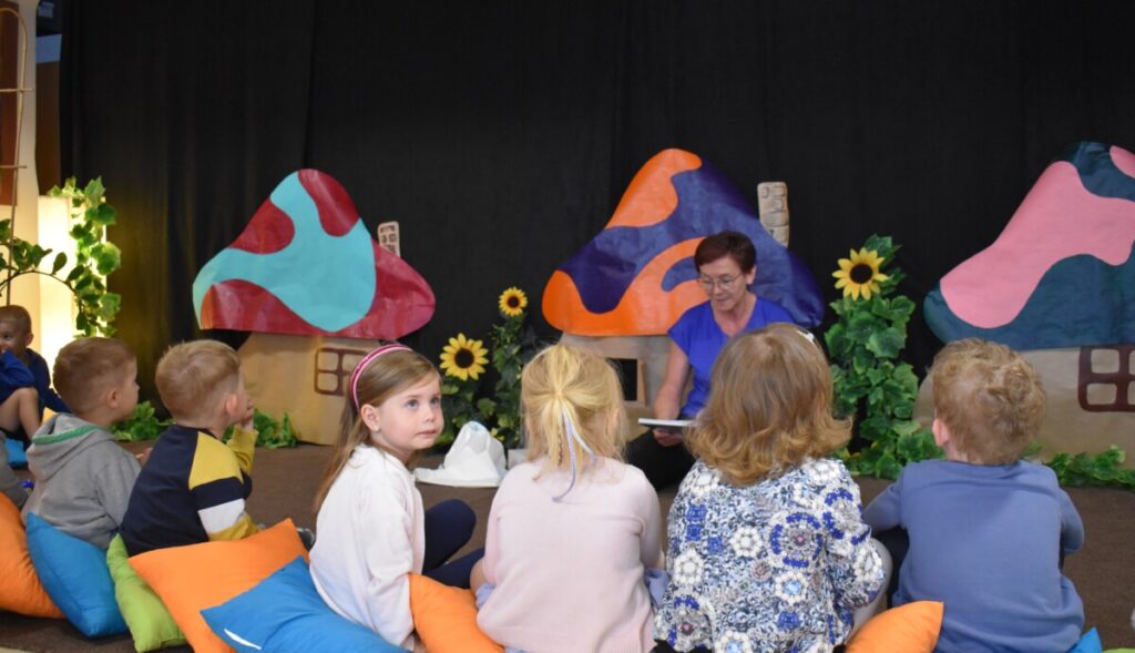 Dzieci siedzą na wykładzinie i kolorowych poduszkach słuchają pani która czyta im książkę, w tle kolorowe chatki smerfów