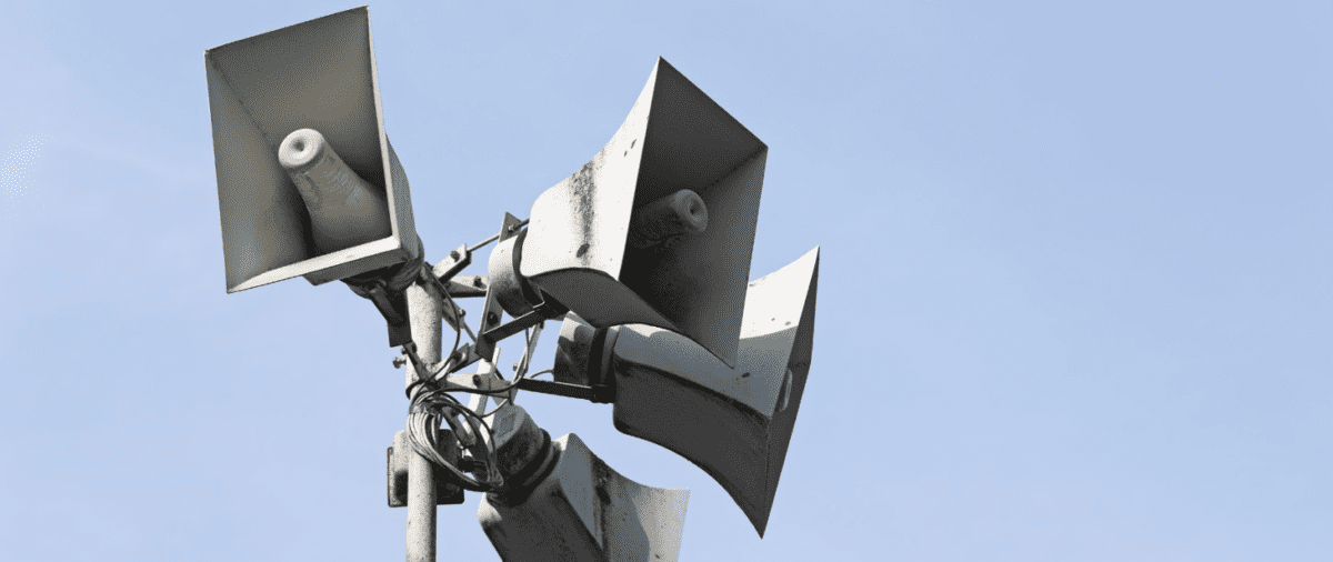 Cztery metalowe megafony na maszcie skierowane w różnych kierunkach. Jasne niebo jako tło. Srebrzysta powierzchnia z efektem patyny.