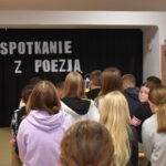 DSC_0372 Dzieci siedzą na ławkach, przed nimi stoi kobieta. W tle czarna kotara na niej napis Spotkanie z poezją