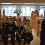 DSC_0414 Na ławkach siedzi grupa dzieci i słucha kobiety, która stoi przed nimi. W tle regały z książkami