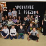 DSC_0418 Grupa dzieci wraz z dwoma paniami stoi na tle kotary z napisem Spotkanie z poezją. W tle widać regały biblioteczne z książkami