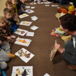 Grupa przedszkolaków i dwie panie pozują do zdjęcia. Dzieci w rękach trzymają kolorowe misie. W tle czarna kotara z białym napisem i gwiazdkami, przed nimi leżą kolorowe poduszki