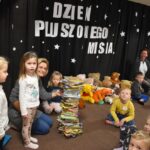 Przedszkolaki siedzą na kolorowych poduszkach i oglądają książki przed nimi leży kilkanaście misiów pluszowych w tle czarna kotara z białym napisem