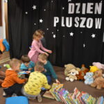 Przedszkolaki siedzą na kolorowych poduszkach i oglądają książki przed nimi leży kilkanaście misiów pluszowych w tle czarna kotara z białym napisem