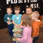 Przedszkolaki siedzą na kolorowych poduszkach i oglądają książki przed nimi leży kilkanaście misiów pluszowych w tle czarna kotara z białym napisem