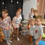 Przedszkolaki siedzą na kolorowych poduszkach i oglądają książki przed nimi leży kilkanaście misiów pluszowych w tle czarna kotara z białym napisem