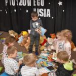 Przedszkolaki siedzą na kolorowych poduszkach i oglądają książki przed nimi leży kilkanaście misiów pluszowych w tle czarna kotara z białym napisem