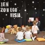 Przedszkolaki siedzą na kolorowych poduszkach i oglądają książki przed nimi leży kilkanaście misiów pluszowych w tle czarna kotara z białym napisem