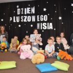 Przedszkolaki siedzą na kolorowych poduszkach i oglądają książki przed nimi leży kilkanaście misiów pluszowych w tle czarna kotara z białym napisem