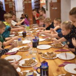 Przy dużym drewnianym stole siedzą dzieci i dekorują pierniczki kolorowymi posypkami, po bu stronach stoją regały z kolorowymi książkami