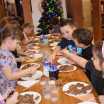 Przy dużym drewnianym stole siedzą dzieci i dekorują pierniczki kolorowymi posypkami, po bu stronach stoją regały z kolorowymi książkami