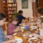 Przy dużym drewnianym stole siedzą dzieci i dekorują pierniczki kolorowymi posypkami, po bu stronach stoją regały z kolorowymi książkami