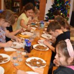 Przy dużym drewnianym stole siedzą dzieci i dekorują pierniczki kolorowymi posypkami, po bu stronach stoją regały z kolorowymi książkami