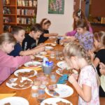 Przy dużym drewnianym stole siedzą dzieci i dekorują pierniczki kolorowymi posypkami, po bu stronach stoją regały z kolorowymi książkami