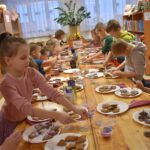 Przy dużym drewnianym stole siedzą dzieci i dekorują pierniczki kolorowymi posypkami, po bu stronach stoją regały z kolorowymi książkami