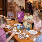 Przy dużym drewnianym stole siedzą dzieci i dekorują pierniczki kolorowymi posypkami, po bu stronach stoją regały z kolorowymi książkami