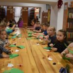 Przy dużym drewnianym stole siedzą dzieci i dekorują pierniczki kolorowymi posypkami, po bu stronach stoją regały z kolorowymi książkami
