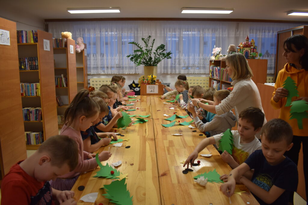 Przy dużym drewnianym stole siedzą dzieci i dekorują pierniczki kolorowymi posypkami, po bu stronach stoją regały z kolorowymi książkami