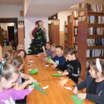 Przy dużym drewnianym stole siedzą dzieci i dekorują pierniczki kolorowymi posypkami, po bu stronach stoją regały z kolorowymi książkami