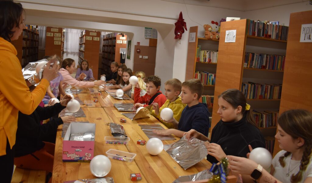 Przy dużym drewnianym stole siedzą dzieci i dekorują pierniczki kolorowymi posypkami, po bu stronach stoją regały z kolorowymi książkami
