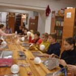 Przy dużym drewnianym stole siedzą dzieci i dekorują pierniczki kolorowymi posypkami, po bu stronach stoją regały z kolorowymi książkami