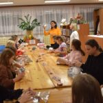 Przy dużym drewnianym stole siedzą dzieci i dekorują pierniczki kolorowymi posypkami, po bu stronach stoją regały z kolorowymi książkami