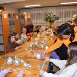 Przy dużym drewnianym stole siedzą dzieci i dekorują pierniczki kolorowymi posypkami, po bu stronach stoją regały z kolorowymi książkami