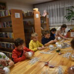 Przy dużym drewnianym stole siedzą dzieci i dekorują pierniczki kolorowymi posypkami, po bu stronach stoją regały z kolorowymi książkami