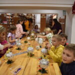 Przy dużym drewnianym stole siedzą dzieci i dekorują pierniczki kolorowymi posypkami, po bu stronach stoją regały z kolorowymi książkami