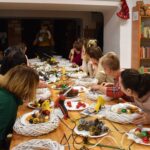 347 Przy drewnianym długim stole siedzą panie i słuchają instruktora. W tle regały biblioteczne z książkami, na końcu stoi kobieta i trzyma w ręku wianek ozdobny na drzwi.