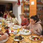 353 Kilkanaście kobiet siedzi przy stole na którym leżą białe wianki ratanowe, kolorowe bombki, szyszki, orzechy, na końcu stołu stoi instruktor i objaśnia zasady robienia dekoracji. W tle jasne drewniane regały z kolorowymi książkami.