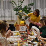 370 Na tle okna stoi kobieta i pokazuje wianek z ozdobami, reszta pań jej słucha. Na stole przy którym siedzą leżą wianki ratanowe, ozdoby, szyszki, bombki.