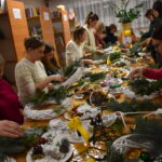 375 Na pierwszym planie stół na którym leży dużo ozdób , świerku, bombek, szyszki. Kobiety dekorują nimi ratanowe wianki. W tle regały z książkami.