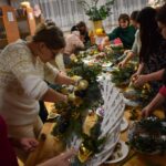 381 Na pierwszym planie wianek ratanowy ozdobiony bombkami, świerkiem, pomarańczami. W tle panie ozdabiają swoje wianki..